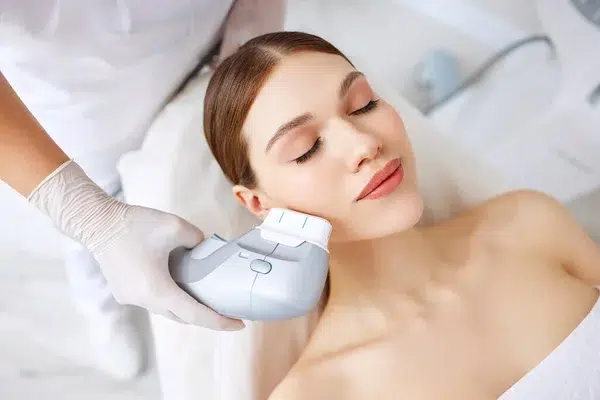 Woman in protective glasses and mask using laser for hair removal on face of client in beauty salon